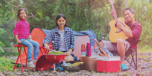 A family of three enjoys camping outdoors. A man plays guitar, while two children sit by a tent with camping gear and food around them. They all appear happy and relaxed in a wooded area.