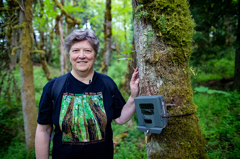 A person with short gray hair stands in a lush green forest, smiling at the camera. They wear a black T-shirt with trees on it and rest their hand on a tree with a mounted camera or sensor device attached to the trunk.