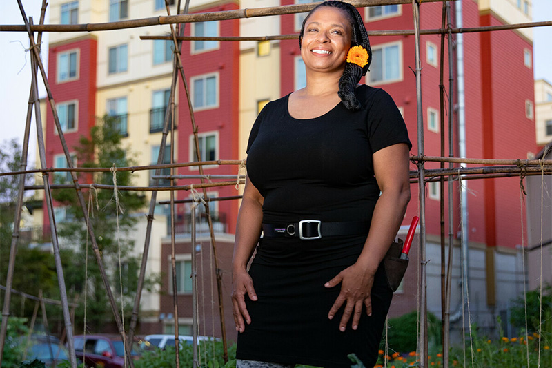 A woman wearing a black dress and tool belt stands and smiles in a garden with wooden supports, with a modern red and beige apartment building in the background. She has a yellow flower in her braided hair.