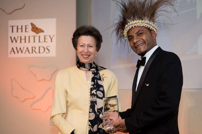 A woman in a yellow jacket stands next to a man in a tuxedo and feathered headdress, who is holding a Whitley Awards trophy. Both are smiling in front of a Whitley Awards sign with butterfly motifs.