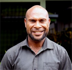 Alfred Kuke. A man with a bald head and short beard is smiling at the camera, wearing a grey collared shirt, standing outdoors with greenery in the blurred background.