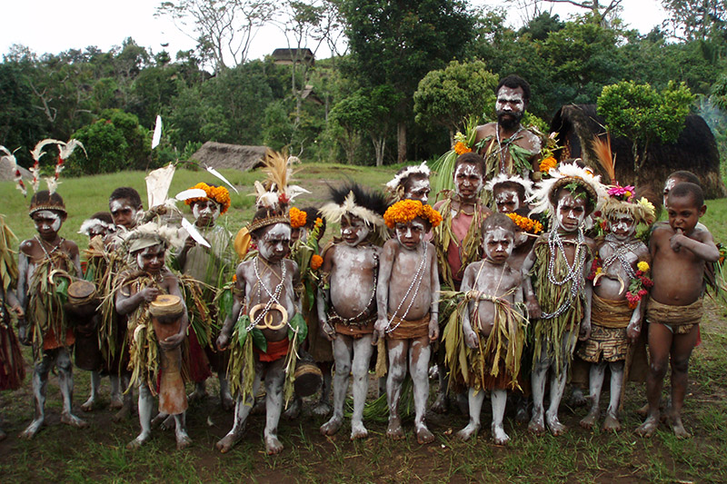 A group of children and adults with painted faces and bodies, wearing traditional grass skirts, jewelry, and headdresses, stand outdoors in a lush green area—reflecting the deep cultural roots and community needs in the YUS region of Papua New Guinea.