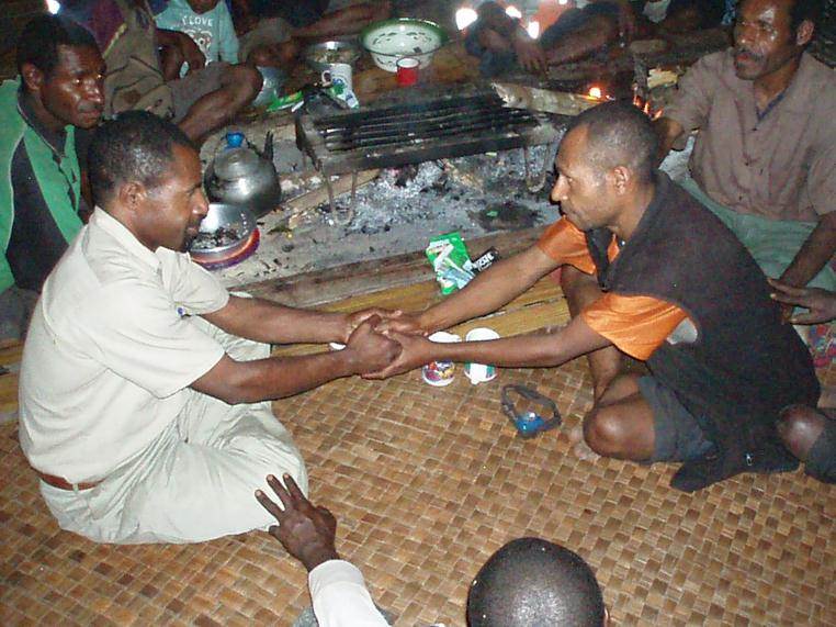 Two men sit cross-legged on a woven mat, holding each other’s hands as others gather in a circle. Cooking pots near a fire suggest a communal gathering focused on providing a sustainable future for the YUS region of Papua New Guinea.