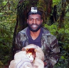 Danny Nane. A man wearing a camo jacket and a head-mounted camera stands in a lush forest, smiling and holding a blanket-wrapped animal with reddish-brown fur and a pale face.