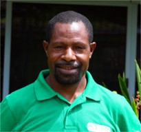 Danny Samandingke, a man wearing a green collared shirt stands outside in front of a building, smiling at the camera. There are green plants partially visible beside him.