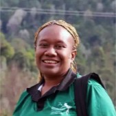Geraldine Tingiran. A woman with braided hair smiles outdoors, wearing a green collared shirt with a blurred forest background.