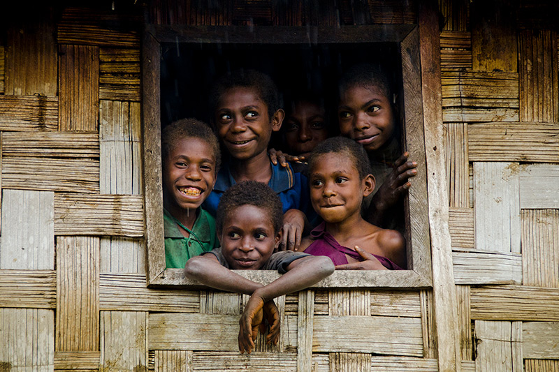 Six children look out and smile from a window of a woven bamboo house.
