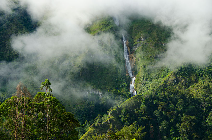 A tall waterfall cascades down a lush green mountainside, partially obscured by mist and clouds, surrounded by dense tropical forest.
