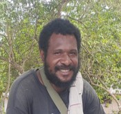 Luther Obert. A man with a beard and curly hair smiles at the camera while sitting outdoors, with green trees in the background. He is wearing a dark shirt and has a bag strap across his shoulder.