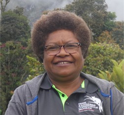 Modi Pontio. A smiling woman with short curly hair, wearing glasses and a dark gray collared shirt with the Tree Kangaroo Conservation Program logo, stands outdoors among trees and greenery.