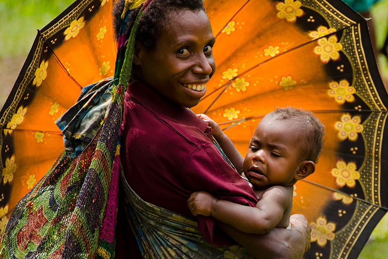 A woman smiles while holding a baby in her arms, standing in front of a large, colorful orange and black umbrella. The scene is filled with bright, natural light.