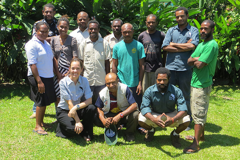 A group of thirteen people, including men and women, stand and kneel together on grass in front of lush green plants, smiling at the camera in a sunny outdoor setting while operating the Tree Kangaroo Conservation Program.
