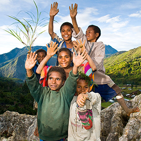 Six smiling children with raised hands stand on rocky ground, with lush green mountains and a village behind them—reflecting the vibrant communities engaged in the Tree Kangaroo Conservation Program under a bright, partly cloudy sky.