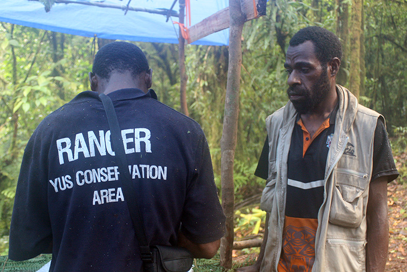 Two men in a forested area; one, helping with managing the YUS region in Papua New Guinea, has "Ranger YUS Conservation Area" on his shirt and stands with his back to the camera; the other faces forward in a vest and orange-striped shirt.