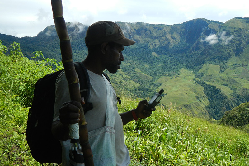 A person holding a walking stick and a device, possibly a GPS, stands on a grassy hillside with a backpack, overlooking the lush, mountainous landscape of the YUS region in Papua New Guinea under a partly cloudy sky.