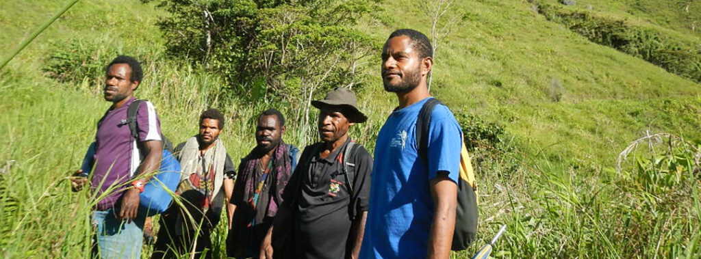 Five men stand together in tall grass on a hillside, surrounded by lush vegetation, with backpacks and outdoor clothing.