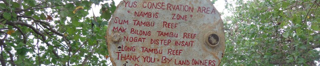 A roughly oval, weathered stone sign with red handwritten text about the YUS Conservation Area surrounded by green leafy branches.