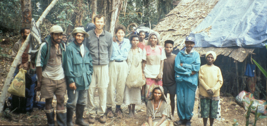 A group of people stand together in front of a rustic shelter in a forested area, reminiscent of gatherings from the history of the Tree Kangaroo Conservation Program. Some are dressed in outdoor clothing as trees and foliage surround the scene, with a few kneeling in front.