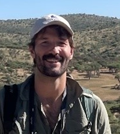 Dr. Samuel Merson, a man with a beard and mustache, wearing a light-colored cap and green shirt, stands outdoors with a hilly, tree-dotted landscape.
