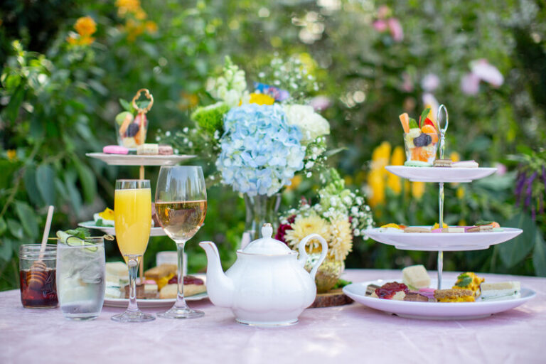 An elegant outdoor table set for tea with a white teapot, glasses of wine, orange juice, and iced drinks, tiered trays of assorted pastries and sandwiches, and a bouquet of blue and yellow flowers in the background.