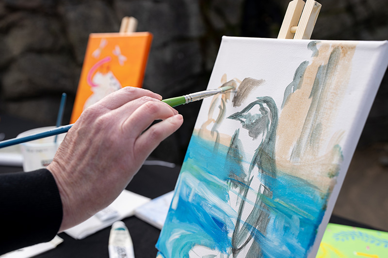 Một bàn tay cầm cọ vẽ đang tô màu trên bức tranh vẽ một chú chim cánh cụt trên nền xanh và nâu; một bức tranh đầy màu sắc khác có thể nhìn thấy ở phía sau.