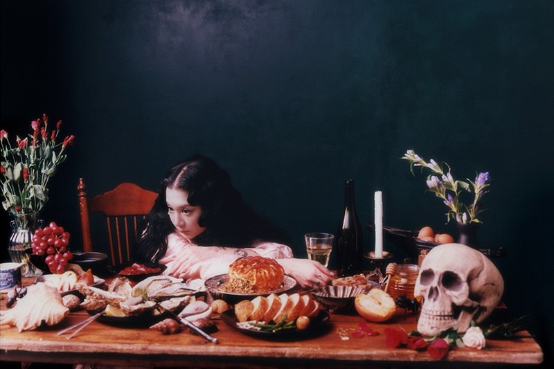 A woman with long dark hair leans on a wooden table covered with food, bread, fruit, wine, flowers, candles, and a human skull, evoking a dark, dramatic, still-life scene against a dark green background.