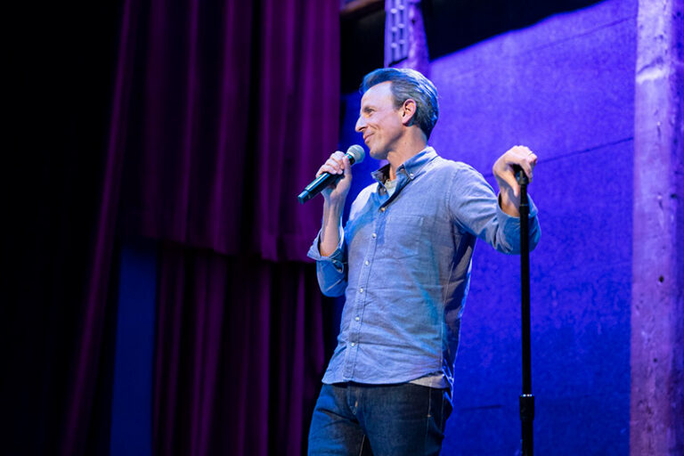 A man in a blue shirt and jeans stands on stage holding a microphone, smiling and speaking. He is lit by stage lights, with a purple curtain and wall in the background.