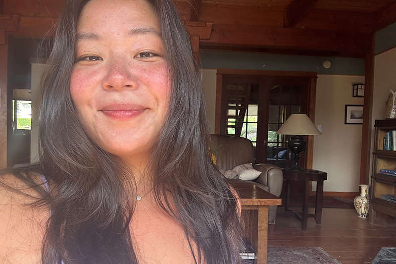 Một người phụ nữ với mái tóc dài màu nâu và khuôn mặt tự nhiên, hơi ửng hồng, mỉm cười với máy ảnh bên trong một căn phòng gỗ ấm cúng với giá sách, đèn và một chiếc ghế bành lớn ở phía sau.