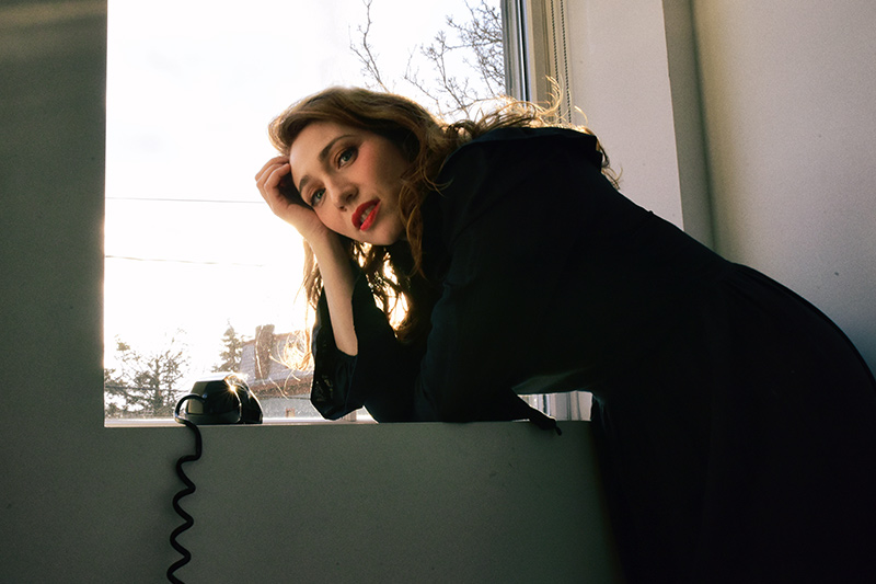A woman with wavy hair and red lipstick leans against a windowsill, resting her head on her hand. She gazes at the camera in soft sunlight, with a black rotary phone on the windowsill beside her.