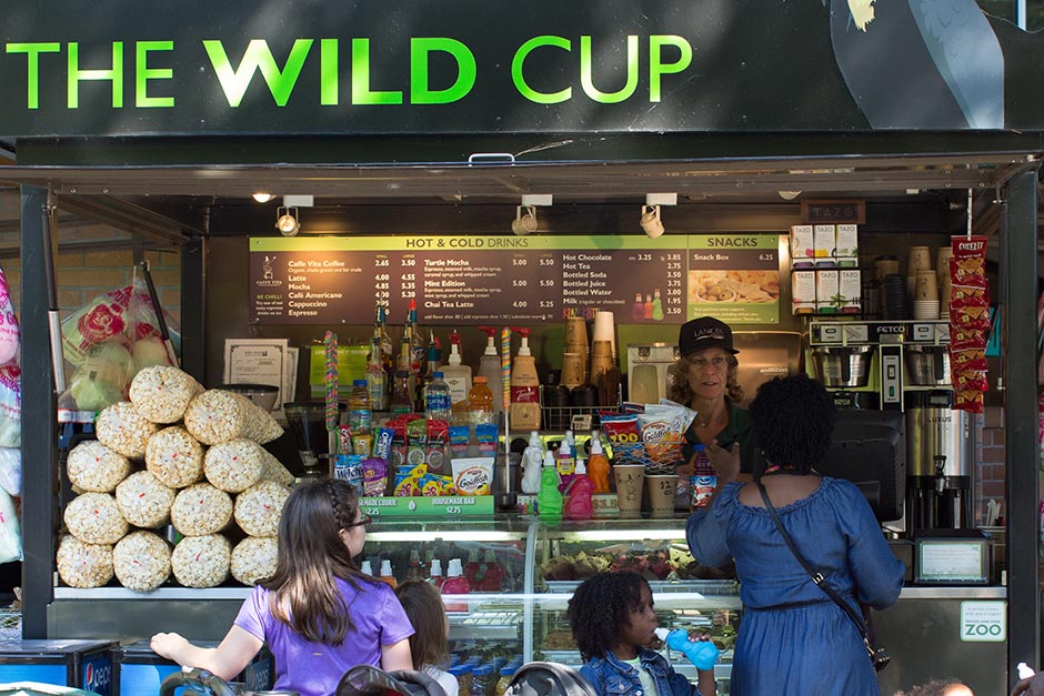 Un puesto de refrigerios llamado The Wild Cup ofrece palomitas, papas fritas, bebidas y bocadillos. Dos mujeres y dos niños están de pie en el mostrador mientras un empleado los atiende. El menú está visible detrás del mostrador.