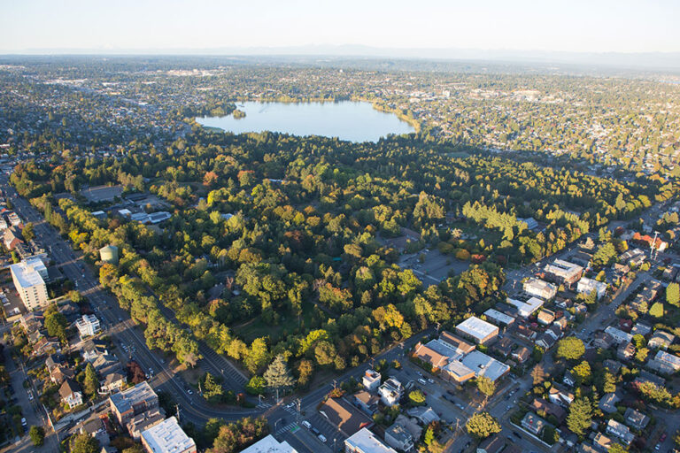 Aerial view of the zoo with dense green trees, surrounded by urban streets and buildings, with a large lake and sprawling cityscape in the background under a clear sky.