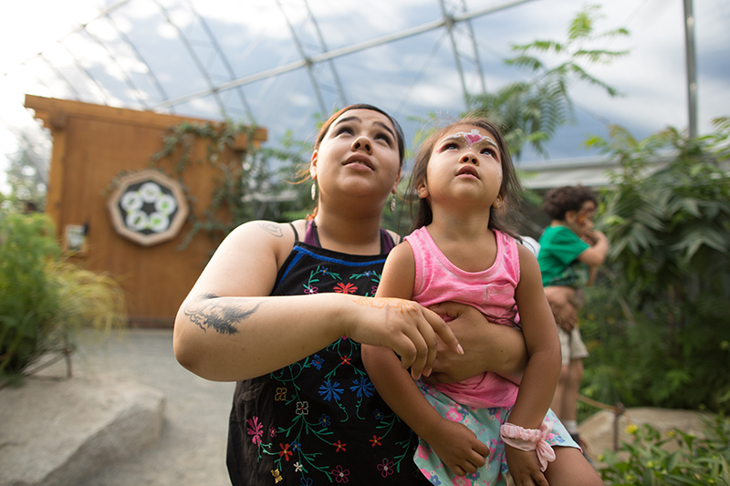 Una mujer sostiene a una niña pequeña mirando hacia arriba en un invernadero, ambas con expresión curiosa. La niña tiene la cara pintada y viste un traje rosa. Al fondo se ven plantas y otra niña.