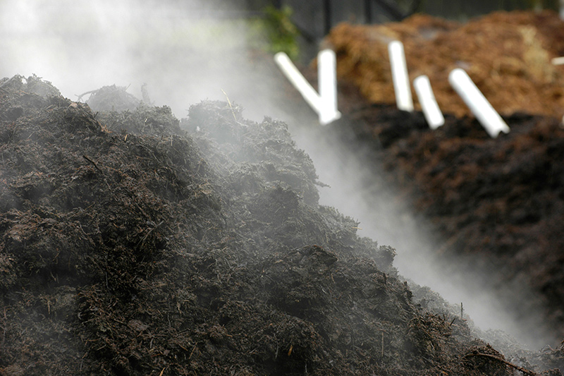 A close-up of a steaming compost pile with visible moisture, with white pipes and more compost in the blurred background.