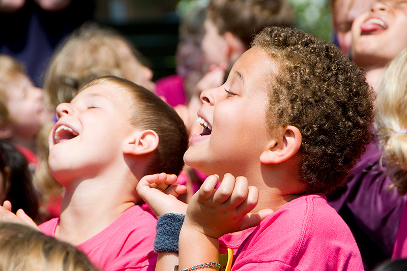 Two young children in pink shirts smile and laugh with their heads tilted back, enjoying a joyful outdoor moment in a group setting.