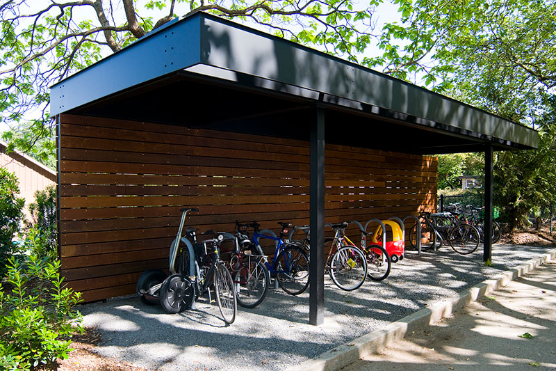 A covered bike shelter with a black metal roof and wooden slat walls, storing several bicycles, a Segway, and a red-and-yellow toy car, surrounded by trees and greenery.