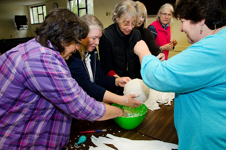 Un grupo de mujeres se reúne alrededor de una mesa, trabajando juntas en una manualidad con un globo de papel maché sobre un tazón verde. Esparcen papel blanco roto y pegamento mientras sonríen y se concentran en la actividad.