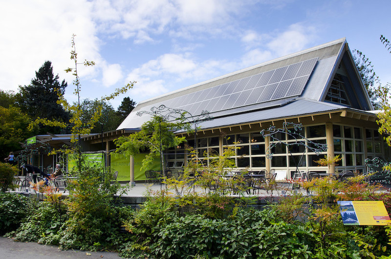A modern building with large windows and solar panels on the roof, surrounded by lush green plants and outdoor seating under a partly cloudy sky.