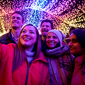 Năm người mỉm cười và tạo dáng chụp ảnh tự sướng dưới một mái vòm được trang trí bằng những ánh đèn rực rỡ sắc màu vào ban đêm. Họ mặc quần áo ấm áp, trông rất vui vẻ và hân hoan.