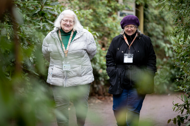Dos mujeres mayores con chaquetas y placas de identificación caminan por un sendero de jardín, sonriendo y charlando. Un exuberante follaje las rodea, creando un ambiente tranquilo al aire libre.