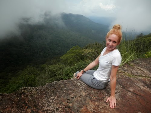 A woman with blonde hair in a bun, wearing a white t-shirt and gray pants, sits on a rocky ledge overlooking a lush green forest and misty mountains in the distance.