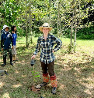 A woman in a plaid shirt, hat, and gaiters smiles while planting a seedling outdoors. Two people stand in the background among trees on a sunny day.