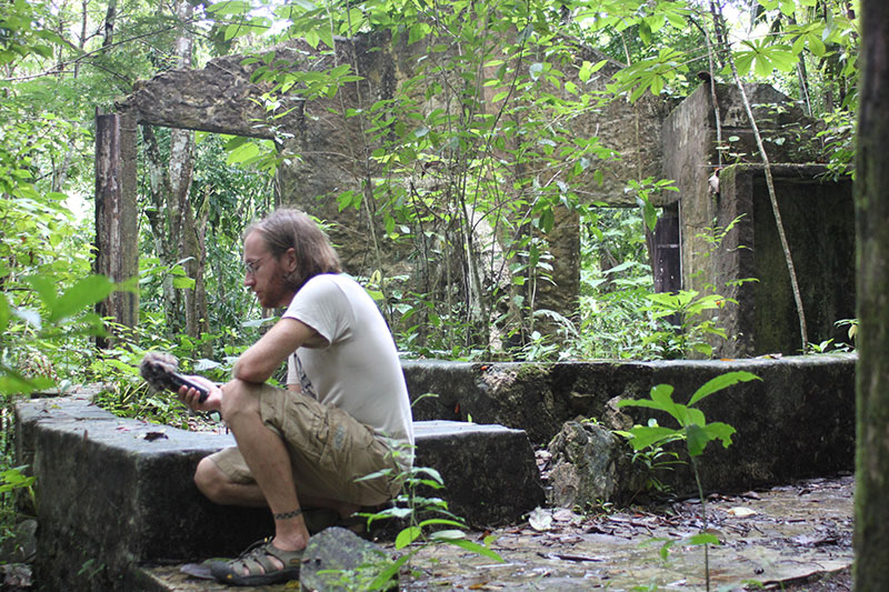 A man with glasses and sandals sits on a stone structure in a lush, overgrown forest, examining an object in his hands. Ruins of a building are visible among the dense greenery.