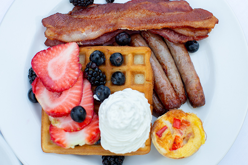 A breakfast plate with waffles topped with strawberries, blueberries, blackberries, and whipped cream, served with bacon strips, sausage links, and a mini egg muffin with red peppers.