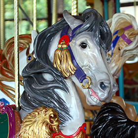 Ảnh cận cảnh một con ngựa gỗ đu quay được sơn vẽ với bờm màu xám, bộ yên cương cầu kỳ và những họa tiết trang trí đầy màu sắc, cùng với một số bộ phận của những con ngựa gỗ đu quay khác ở phía sau.