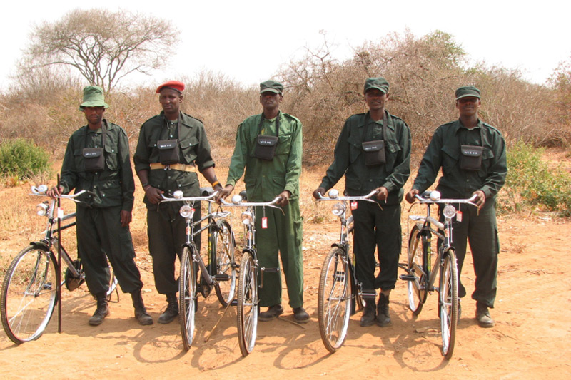 Five uniformed individuals stand outdoors on dirt ground, each holding a bicycle. They wear dark green clothing and caps, with some wearing red or green berets. Sparse trees and dry bushes are visible in the background.