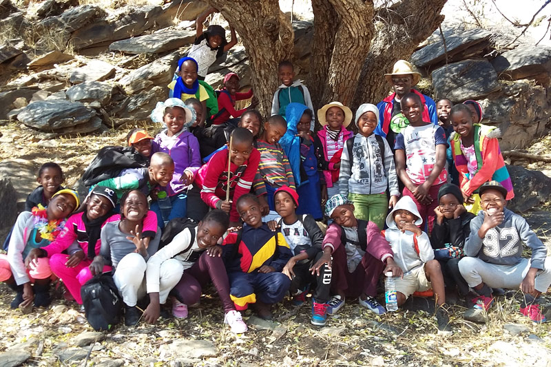 A group of smiling children pose together outdoors under a large tree, wearing colorful clothes and hats, with rocky terrain in the background.