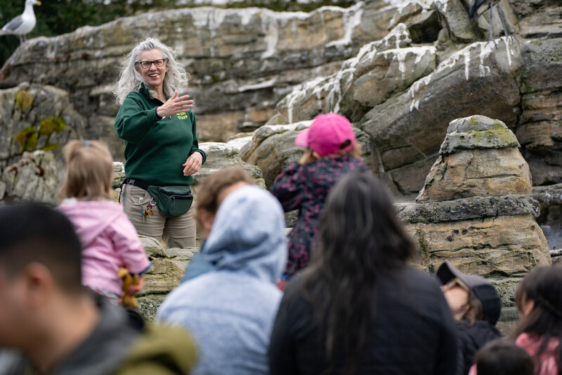 A woman in a green sweater speaks to a group of people outdoors in front of rocky terrain. The listeners, including children and adults in jackets and hats, face her as she gestures with one hand.