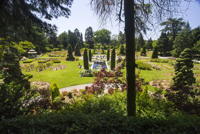 A lush, formal garden with manicured lawns, blooming flower beds, sculpted topiary trees, pathways, and a central reflecting pool, all surrounded by tall trees under a clear blue sky.