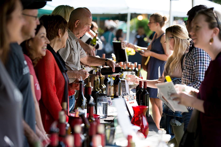 People interact and sample wine at an outdoor market or festival. Bottles of wine line the tables as customers and vendors smile and converse in a lively, sunlit setting.