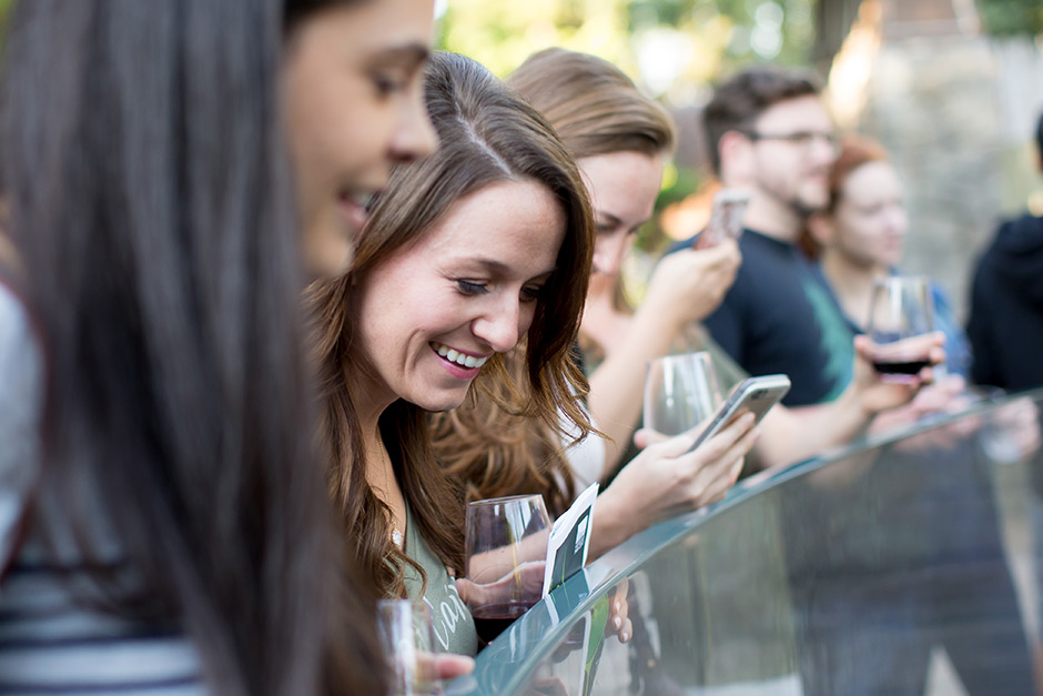 A group of people stand outdoors by a glass railing, smiling and socializing. One woman in focus holds a glass of wine and looks at a phone, while others chat and enjoy drinks in the background.